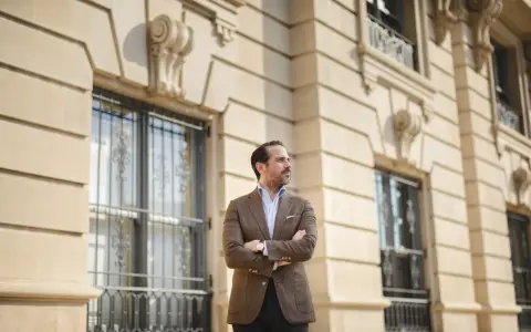 A man in a stylish brown sport coat outfit stands with arms crossed in front of a classic building.