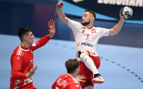 Gracz w piłkę ręczną w skoku, z piłką w dłoni, celuje do bramki. Jego przeciwnik próbuje go zatrzymać.