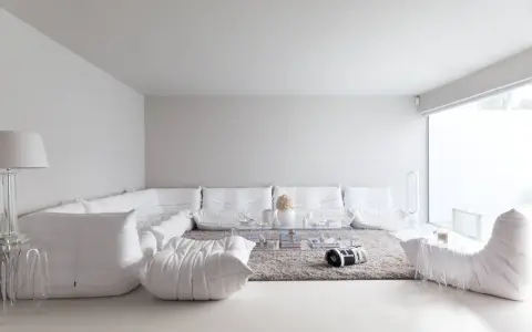 Soggiorno minimalista con divano componibile bianco, tappeto grigio e tavolino trasparente. Le tonalità di bianco per pareti creano un'atmosfera eterea.