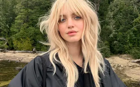 Blonde Frau mit lässigem Wolf Cut stylen, blauen Augen und einem schwarzen Jackett vor einer Waldkulisse.
