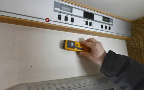 Hand håller fuktmätare mot vägg för fukttest protokoll.