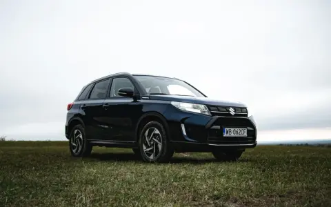 Ciemnoniebieski Suzuki Vitara Hybrid na tle zachmurzonego nieba, zaparkowany na trawiastym wzgórzu.