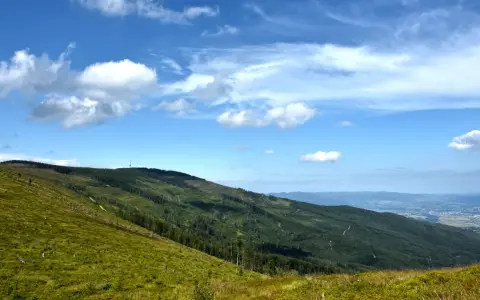 Zielone, trawiaste zbocze schodzi ku lasom i miastu w oddali. W tle widać kolejne pasma górskie i niebo z chmurami. To malownicze **beskidy szczyty**.