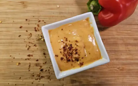 Cremige Chipotle-Sauce in einer weißen Schale, bestreut mit Chiliflocken. Daneben liegt eine rote Paprika auf einem Holzbrett.
