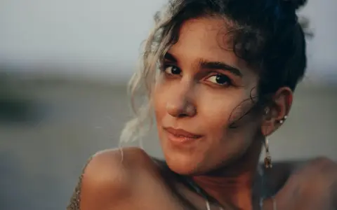 Photos de portrait d'une jeune femme aux cheveux bouclés, le sable sur son épaule, regardant la caméra avec un léger sourire.