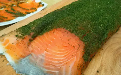 Un gran filete de salmón marinado casero cubierto de eneldo fresco. En el fondo, finas lonchas de salmón ya preparadas.