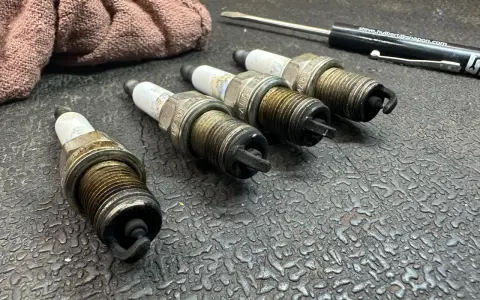 Four used spark plugs lie on a textured surface next to a rag and a screwdriver. This image might make you wonder how many spark plugs a car has.