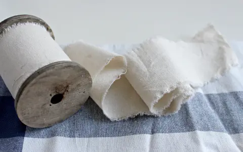 Un vecchio rocchetto di legno con tessuto di cotone pettinato arrotolato. Il tessuto è grezzo e sfrangiato, adagiato su una tovaglia a quadri blu e bianchi.