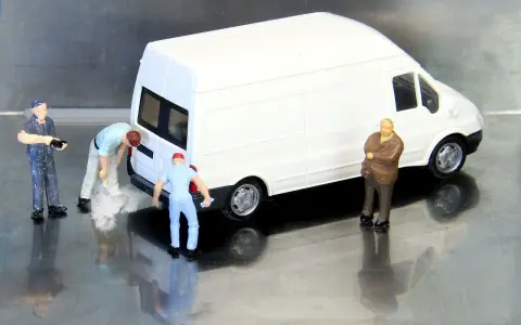 Miniaturowi pracownicy zajmują się białym busem budowlanym. Jeden z nich czyści tył pojazdu, inny trzyma narzędzie, a trzeci obserwuje.