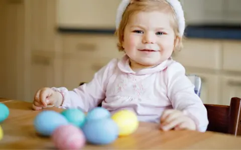 Kleines Mädchen mit Hasenohren und bunten Ostereiern. Sie sitzt am Tisch und schaut neugierig auf das **Baby Ei**.