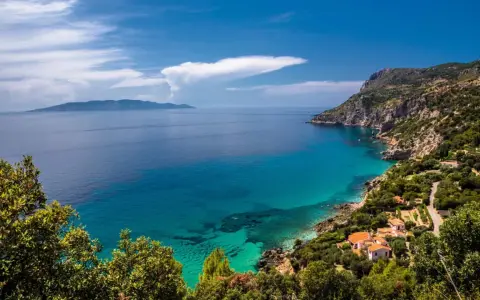 Turquoise water laps a rocky Tuscan coast with villas nestled among trees, under a blue sky with clouds.