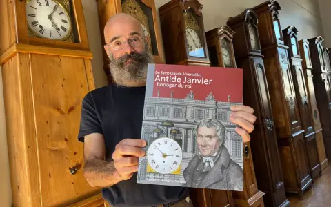 Un homme barbu présente un livre sur Antide Janvier, horloger du roi, devant une rangée de grandes horloges anciennes.