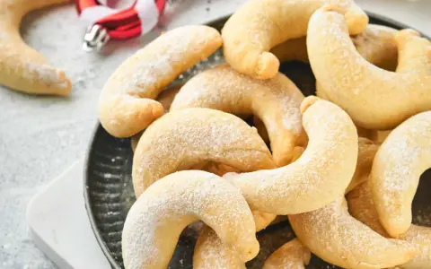 Ein Haufen goldbrauner Vanillekipferl, bestäubt mit Puderzucker, liegt auf einem dunklen Teller. Im Hintergrund sind weihnachtliche Zuckerstangen zu sehen.