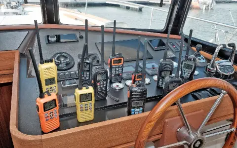 Plusieurs talkies-walkies sur un tableau de bord de bateau. Ils sont de différentes couleurs (orange, jaune, noir) et affichent des canaux. Une discussion sur la différence entre UHF et VHF pourrait être animée avec ces appareils.