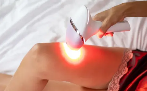 Dłoń trzyma depilator laserowy nad udem, emitując czerwone światło. Czy depilacja laserowa jest szkodliwa?