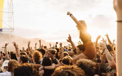 Menschen jubeln auf einem Festival in Rostock. Eine Frau sitzt auf den Schultern eines anderen Besuchers, während die Sonne untergeht.