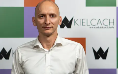Łysy mężczyzna w białej koszuli na tle kolorowej ściany z logo "wKielcach".