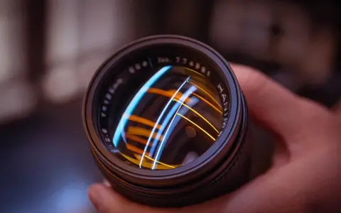 Primer plano de un objetivo fotográfico con reflejos de luces azules y naranjas.