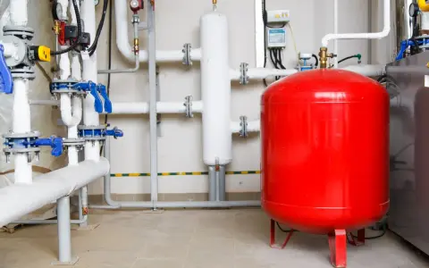 Un sistema de tuberías con válvulas azules y un gran tanque rojo, que es un vaso de expansión, en una sala técnica.