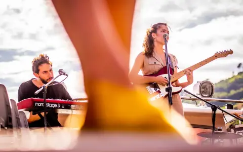 Maria y Feu, una banda en vivo, tocan en un escenario al aire libre. Ella canta y toca la guitarra, él en los teclados.