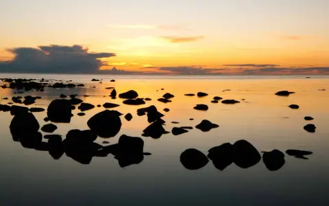 Solnedgång över havet vid en stenig strand på ar Gotland. Mörka stenar speglas i det lugna vattnet under en himmel i orange och gult.