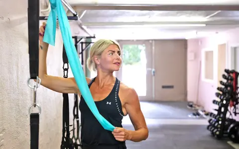 Kvinna med blont hår stretchar axlar med ett turkost träningsband i ett gym.