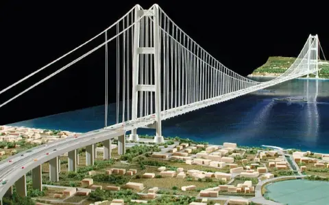 Maqueta de un puente colgante con pilares y cables que forman **puentes triangulados**, cruzando un cuerpo de agua y conectando dos orillas con una ciudad en miniatura.