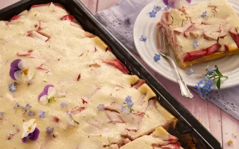 Saftiger Rhabarberkuchen mit Quark, dekoriert mit Veilchen und Vergissmeinnicht. Ein Stück ist auf einem Teller angerichtet.