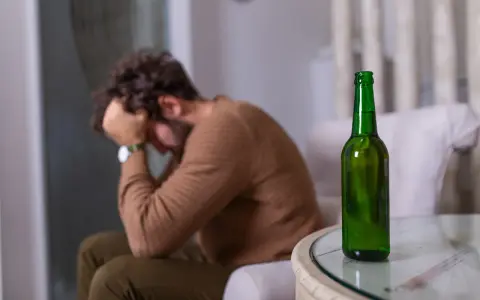 Mężczyzna siedzi z głową w dłoniach, obok stoi zielona butelka. Czuje duszności po alkoholu.