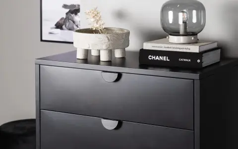 Svart sovrum med en mörk byrå. På byrån står en kruka med torkad växt, en lampa och böcker, bland annat "CHANEL CATWALK".
