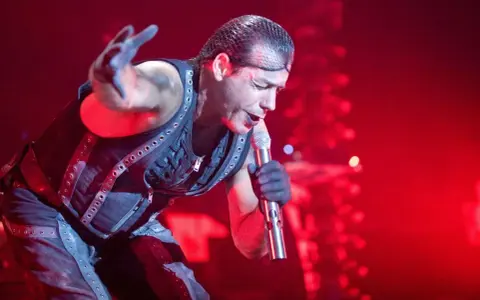 Till Lindemann, chanteur du groupe metal allemand Rammstein, en pleine performance sur scène, micro à la main, sous un éclairage rouge intense.