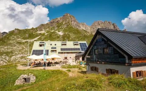 Die Buchel Alpe, eine Berghütte mit Solaranlagen, liegt vor einer beeindruckenden Bergkulisse. Gäste sitzen draußen unter Sonnenschirmen.