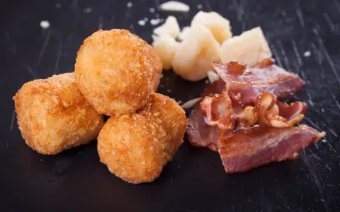Croquetas doradas y crujientes con trozos de bacon y queso sobre fondo oscuro.
