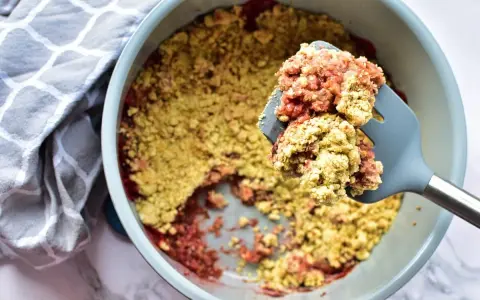 Ein köstlicher Erdbeer Rhabarber Crumble wird mit einem Löffel aus einer Auflaufform gehoben. Daneben liegt ein gemustertes Küchentuch.