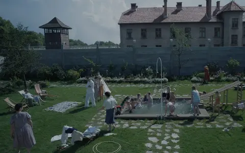 Scena da film strani: bambini giocano in piscina, adulti sorvegliano, mentre sullo sfondo incombono torri di guardia e un edificio imponente.