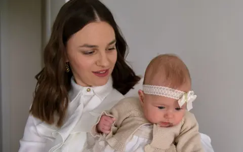 Elegancka mama z niemowlakiem. Inspiracja, jak ubrać się na chrzciny jako gość: biała koszula, subtelne dodatki.