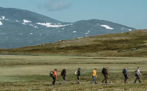 Vandring i fjällandskap med snöklädda toppar, en grupp vandrare med ryggsäckar rör sig genom gräs och ljung mot Helags Sylarna.