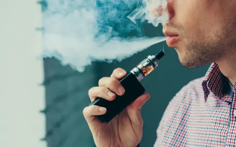 Hombre vapeando, exhala humo blanco. El vapear sube la tensión, cuidado con tu salud.
