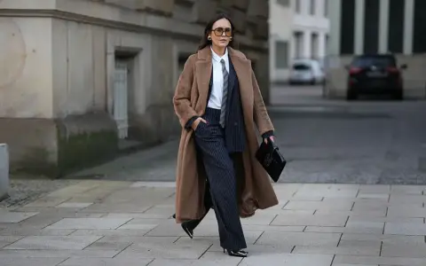 Chica tomboy con abrigo largo marrón, traje de raya diplomática y bolso negro.