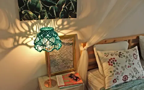 Oświetlona zielona, pleciona lampa tworzy cień na ścianie. Obok łóżko z poduszką. Inspiracja, jak odnowić stary abażur.
