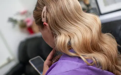 Blond włosy ułożone w hollywoodzkie fale na bok, ozdobione błyszczącą spinką. Kobieta w fioletowym szlafroku przegląda telefon.