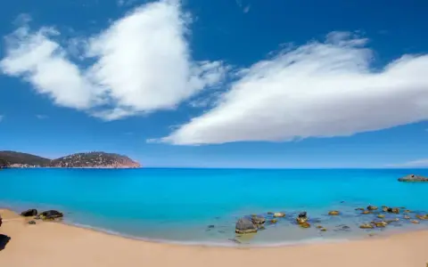 Traumhafter ibiza strand mit türkisblauem Wasser, goldenem Sand und einer felsigen Küste unter einem strahlend blauen Himmel mit weißen Wolken.
