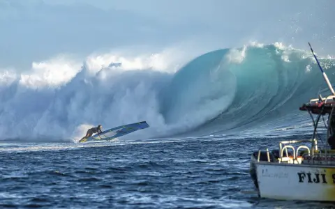 Sarah Hauser surft auf einer riesigen Welle, die sich hinter ihr bricht. Ein Boot mit der Aufschrift "Fiji S" liegt im Vordergrund.