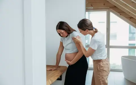 Eine schwangere Frau stützt sich auf einen Tisch, während eine andere Frau ihr hilft. Es ist Zeit, ins Krankenhaus zu fahren.