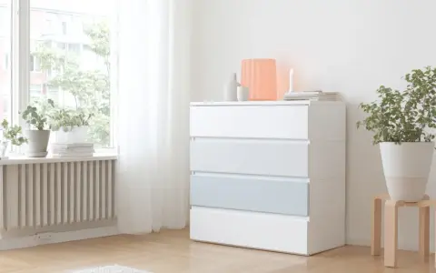 Eine weiße MALM Kommode mit vier Schubladen, eine davon hellblau, steht neben einem Fenster. Darauf sind Deko-Objekte und eine Kerze.