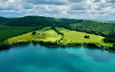Acque turchesi del lago di Martignano lambiscono dolci colline verdi, con un casale isolato tra i campi.