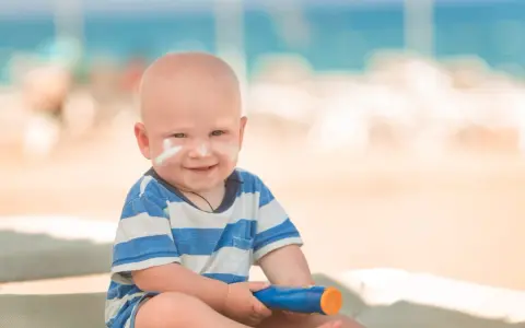 Un bébé souriant avec de la crème solaire sur le visage, tenant un tube de crème solaire. Idéal pour savoir crème solaire bébé à partir de quel âge.