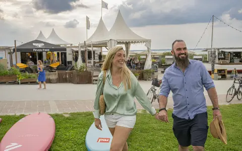 Glückliches Paar spaziert am **Stein Ostsee Strand** entlang, vorbei an SUP-Boards und Strandbuden.