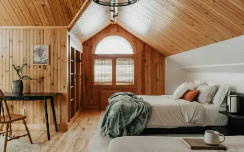 Dormitorio con paredes y techo de madera, cama con mantas y almohadas, ventana arqueada y escritorio.