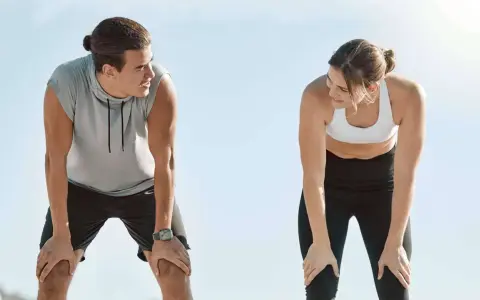 Zwei Läufer erholen sich nach einem anstrengenden 10 km Lauf. Sie schauen sich an, die Hände auf den Knien, erschöpft, aber zufrieden.